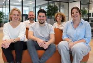 Fünf lächelnde Teammitglieder sitzen auf einem orangefarbenen Sofa in einem modernen, hellen Büro mit Glaswänden.