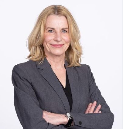 Porträt einer lächelnden Frau mit blondem Haar im grauen Blazer, die die Arme verschränkt vor hellem Hintergrund steht